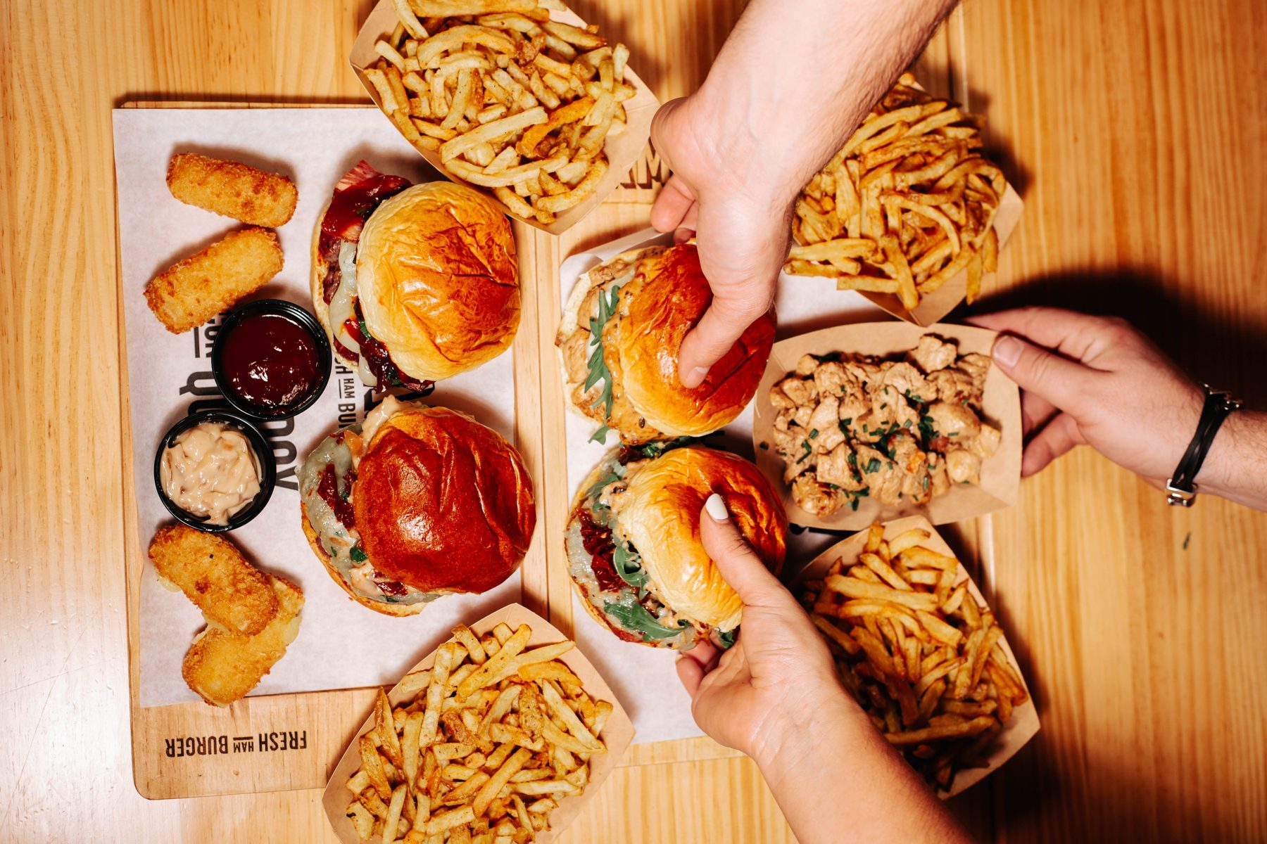 Hamburguesas, patatas y salsas en cenital mientras varias manos las cogen de la mesa